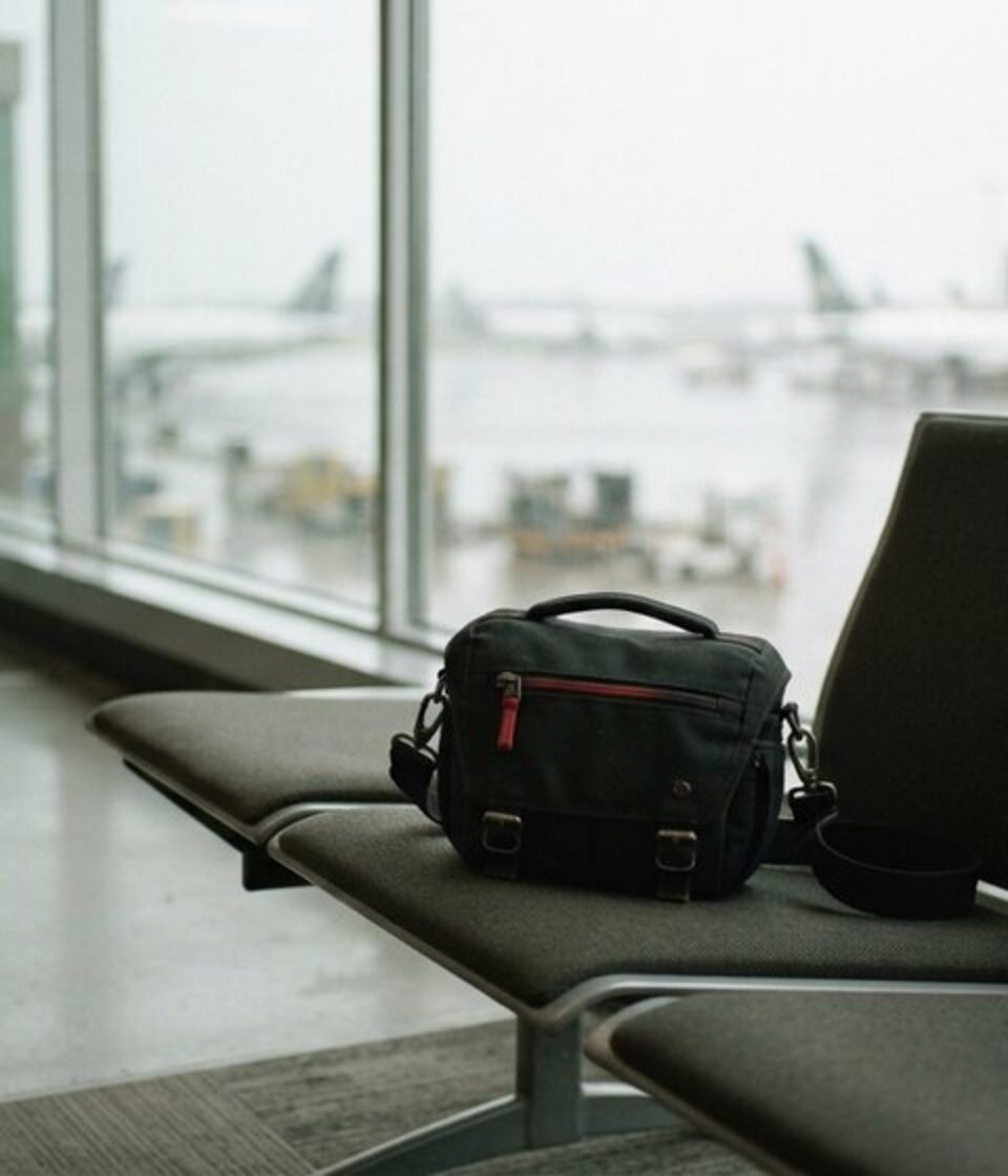 A black backpack left on an airport bench in front of a window overlooking the apron.